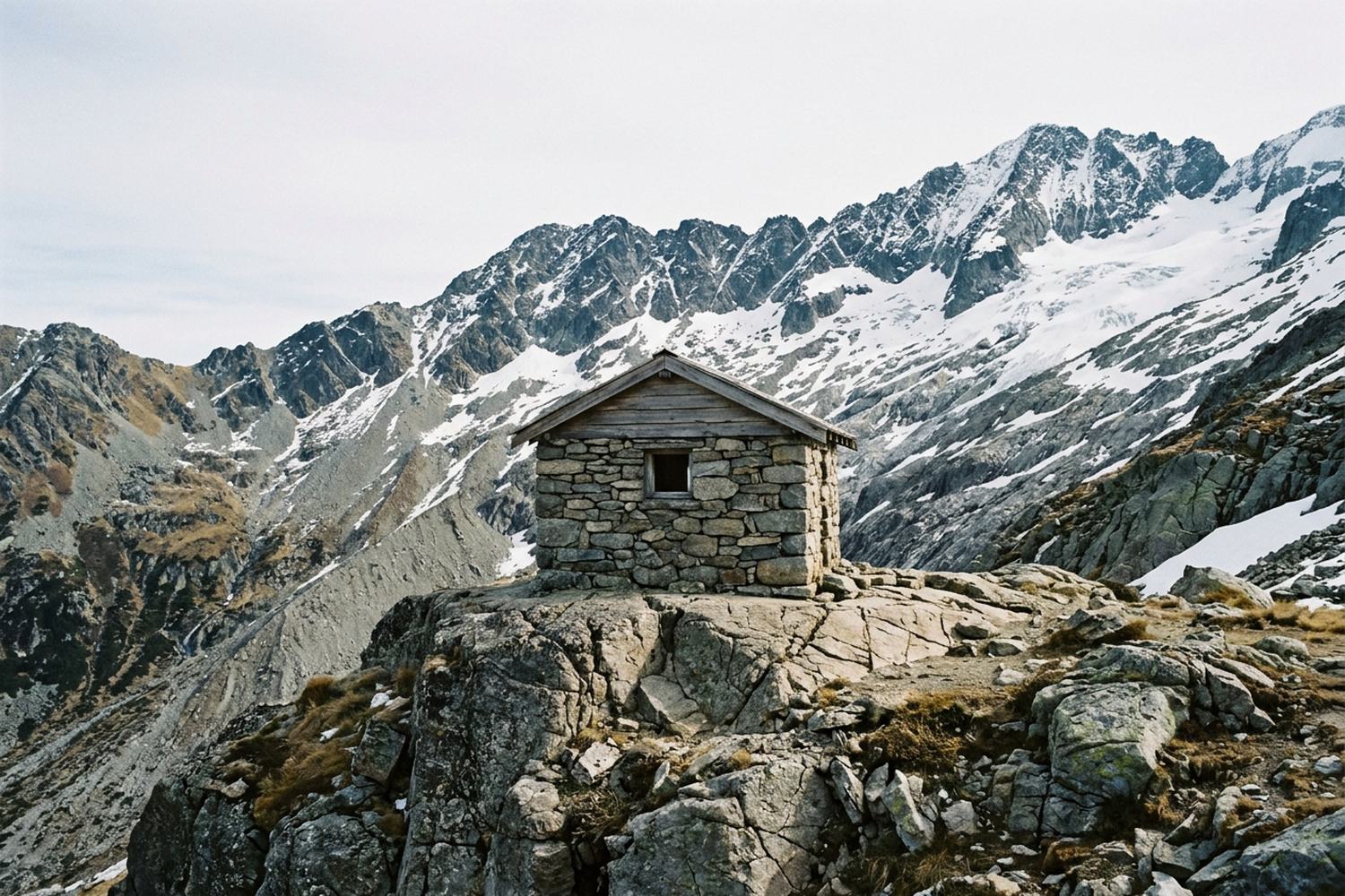 A stone building on a rock ledge below a snow rimmed ridge.